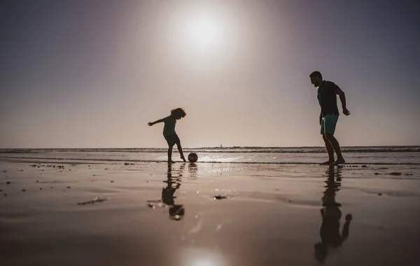 父と息子はビーチでサッカーやサッカーをしたり、日没のシルエットをしたりします。屋外で楽しむお父さんと子供。ライフスタイルと家族の休暇、幸福の男性の概念. — ストック写真