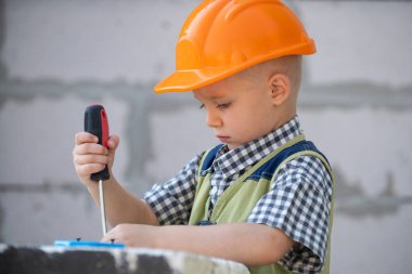 Çocuk tornavidayla cıvata çeviriyor. Tamir aletli bir çocuk tamircisi. İnşaat alanında kasklı çocuk ve içki tulumu var. Küçük işçi mühendisi. Evi onarın..