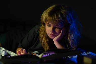 Küçük bir çocuk karanlık evde kitap okuyor. Çocukların yüzü gece lambasıyla kaplıdır. Hayal gücü ya da öğrenme kavramı.