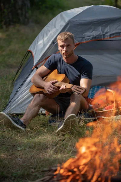 Homem bonito tocando guitarra na floresta com fogueira. Homem romântico acampando ao ar livre e sentado perto da tenda. Homem com guitarra no acampamento. Canções de fogueira. Guitarrista acústico no campo. — Fotografia de Stock