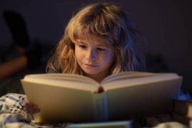 Yatakta uzanmış kitap okuyan küçük sevimli çocuğun yakın plan portresi. Çocuk yatmadan önce kitap okuyor..