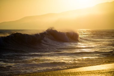 Dalgalar sıçrıyor. Altın gökyüzü ve deniz suyu. Günbatımında deniz manzarası ve tropikal plaj ya da tatil ve tatil için gün doğumu. Okyanus dalgaları arka planı. Suda güneş, kumsalda kum..