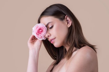 Yüzünde pembe bir gül olan güzel bir kızın yakın plan portresi. Güzel kadın yüzü ve çiçek. Yakın plan portre, cilt bakımı, makyaj.