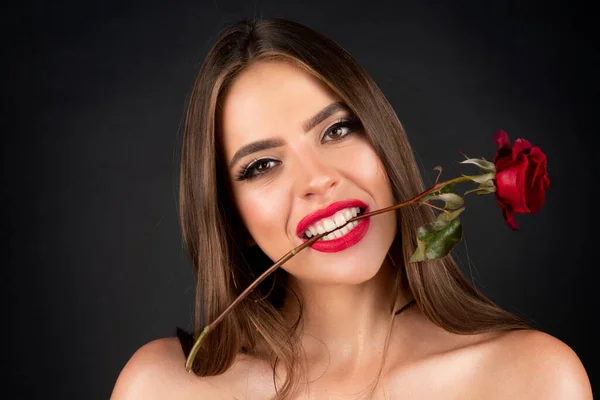 Kobieta z czerwonymi ustami i kwiatem róży. Kwiaty róży w ustach. Piękny portret mody na tle studia. Uwodzicielska kobieta trzyma czerwoną różę z zębami na czarnym, izolowanym tle. — Zdjęcie stockowe