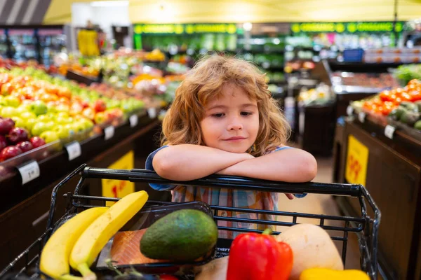 Barnpojken handlar i en stormarknad. Liten söt pojke med kundvagn full av färska ekologiska grönsaker och frukter i mataffär eller stormarknad. — Stockfoto