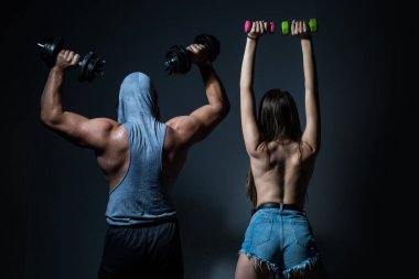 Bir çift formda vücut geliştirici, kaslı bir adam ve dambılı güzel seksi bir kız. Spor çifti. Çıplak vücutlu kaslı bir adam, koyu arkaplanda halterli bir kadın, sırt..