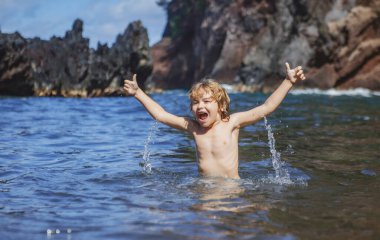 Şaşkın çocuk oynuyor ve denize su sıçrıyor. Çocuk dışarıda eğleniyor. Yaz tatili ve sağlıklı aile hayatı konsepti. Küçük çocuk elini kaldırıp suya su damlatıyor..