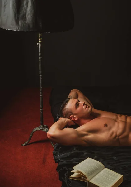 寝室で読書中のセクシーな男。ベッドの中で裸のマシン。セックスとリラクゼーションの概念。本好き. — ストック写真