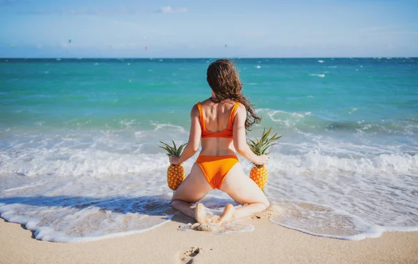 Belle fille modèle sexy en bikini au bord de la mer de l'île tropicale. Voyage vacances d'été. Femme sexy tenant des ananas, vue sur le dos. — Photo