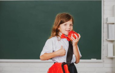 İlkokulda sınıfta kırmızı elma yiyen neşeli bir kızın portresi. Çocukların eğitimi ve bilgisi. Öğrenci çocukları.