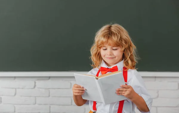 School jongen leest een boek in de klas. Leuk kind op school. Kind leert in de klas op de achtergrond van schoolbord. Terug naar school, eerste schooldag. — Stockfoto