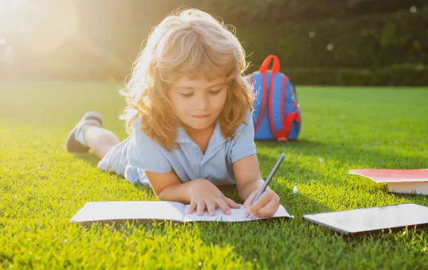 Leuke kinderjongen met boeken met potlood die buiten op notitieboekje schrijven. Zomerkamp. Kinderen leren en onderwijs concept. Zomer vakantie huiswerk. Buitenschoolse kleuterschool. — Stockfoto