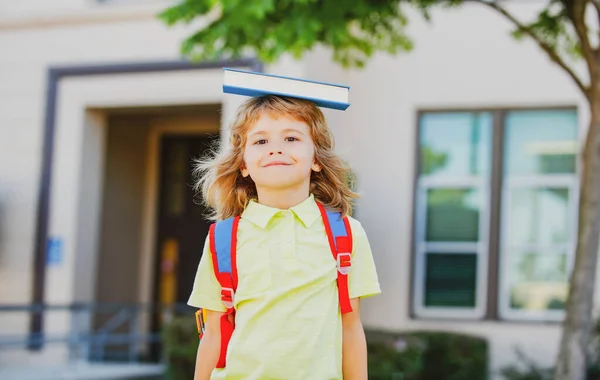 Возвращайся в школу. Забавный мальчик в очках рядом со школой. Ребенок из начальной школы с книгой и сумкой. Образование. Парень с книгой. — стоковое фото