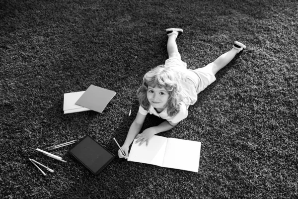 Miúdo lendo livro de interesse e escrevendo no caderno no jardim. Divertimento de verão. Rapaz bonito deitado na grama lendo um livro de crianças. — Fotografia de Stock