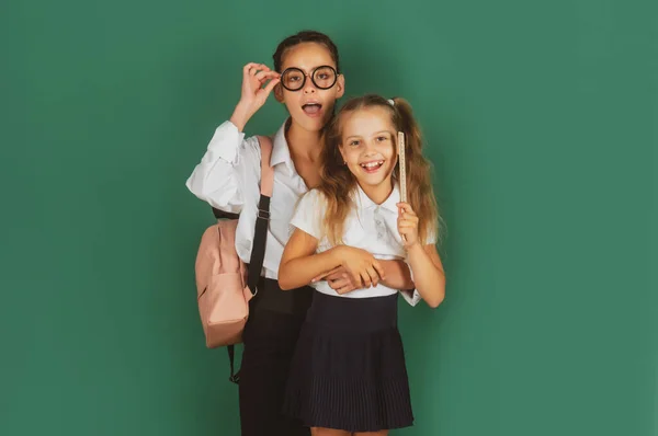 Dia do conhecimento, sorrindo engraçadas meninas colegiais irmãs com mochila segurar livros em quadro-negro verde. Conceito de estilo de vida infantil. Educação na escola. — Fotografia de Stock