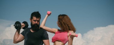 Dışarıda dambılları olan sağlıklı bir çift atletik erkek ve kadın. Dışarıda spor yapan bir çift. Genç fitness çifti, afiş. Spor mankenleri kollarını dambılla şişiriyor..