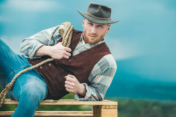 Bauer Cowboy mit Hut. Westliches Leben. Amerikanisches Landmännchenporträt. Landarbeiter auf dem Land auf dem Bauernhof oder auf der Ranch. Cowboy mit Lassoseil auf Himmelshintergrund. — Stockfoto