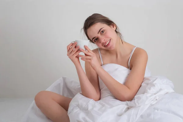 Goeiemorgen. Vrolijke jonge vrouw met kopje koffie of thee in bed in de slaapkamer. Portret van een mooie vrouw in witte pyjama die koffie drinkt. — Stockfoto
