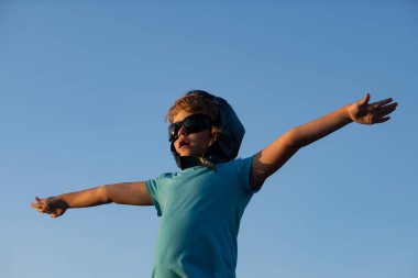 Pilot gözlüklü ve kasklı mutlu çocuk mavi gökyüzü arka planına karşı oynuyor. Güneşli bir yaz gününde gökyüzünde yürüyen sevimli bir çocuk. Çocuklar seyahat hayali kurar..