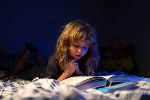 Barn läser en bok i det mörka hemmet. Barnpojken läser en bok som ligger på sängen. Mot kunskap. Pojke med bok läsa berättelse över mörka nära rummet bakgrund. Bokhandelskoncept. — Stockfoto