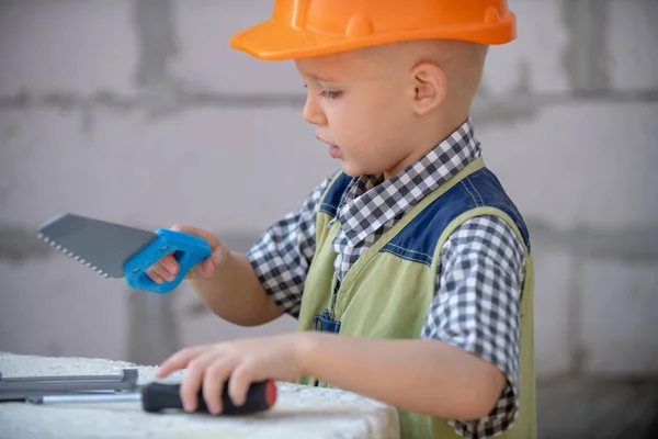Preschool boy trying to saw, Child sawing on a construction site near house wall. Blond child with helmet uses saw. — Stock Photo, Image