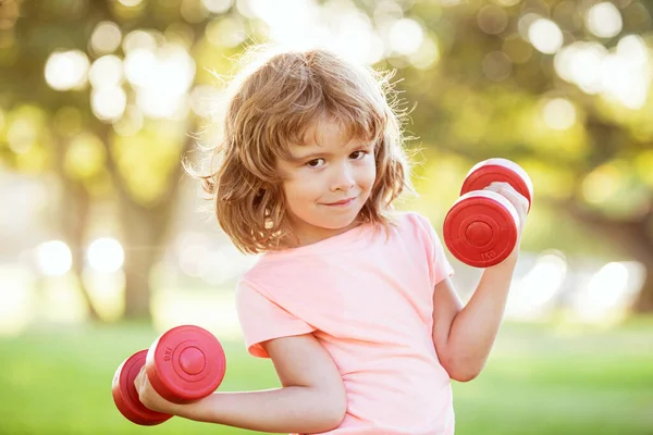 Dambılı şirin bir çocuk. Çocuklar spor ve spor, sağlık ve enerji. Sağlıklı çocuk yaşam tarzı. Neşeli çocuk halterlerle egzersiz yapıyor. Çocuk dambıllarla egzersiz yapıyor. Açık hava çocukları. — Stok fotoğraf