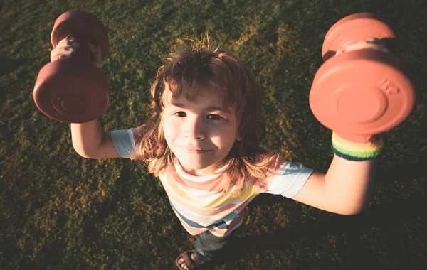 Lustiger Kindersport. Kind mit Hanteln im Park. Starker kleiner Junge. Lustiges Kindergesicht. Entwicklung der Kinder und gesunde starke Bewegung. Weitwinkel. — Stockfoto