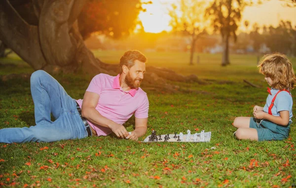 幸せな家庭の屋外。夏の庭でチェスをする父と息子。男の子は親とチェスをする。認知発達。知的子供向けゲーム。賢い子供たち. — ストック写真
