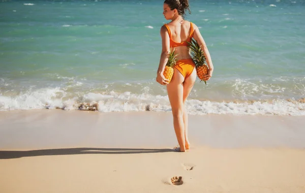 Kvinna med sexiga skinkor i baddräkt. Ung kvinna som håller en ananas på dominikanska eller hawaii stranden. Varm sommarsemester på stranden. — Stockfoto