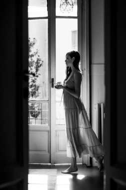 Elinde bir kupa tutuyor, mutlu dans ediyor. Kız gece kıyafeti giymiş. Ev manzaralı balkon. Uzun saçlı güzel bir kız portresi. Sabahın doğuşunda.