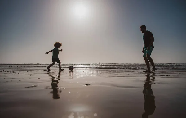 父と息子はビーチでサッカーやサッカーをします。父と子供の屋外で、日没のシルエットを再生します。アクティブファミリーコンセプト. — ストック写真