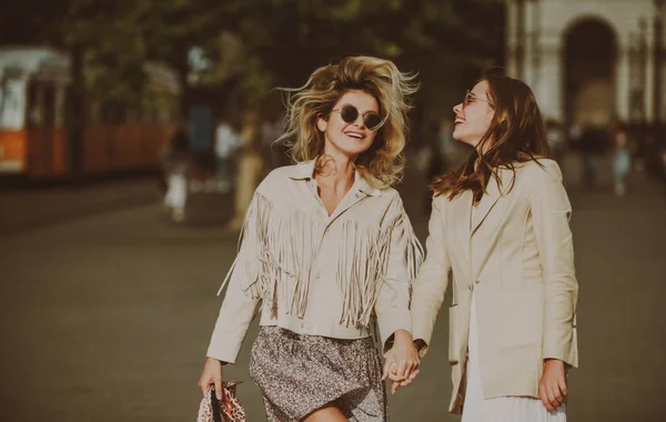 Dos mujeres emocionadas caminando por la calle. Hermosa chica cogida de la mano con un amigo mientras camina por la ciudad. Retrato al aire libre de dos hermanas riendo pasando tiempo libre juntas. — Foto de Stock