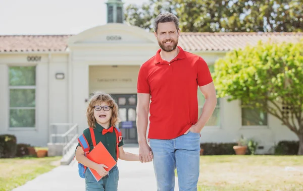 Parent and pupil of primary school go hand in hand. Teacher in t-shirt and cute schoolboy with backpack near school park. — Stock Photo, Image