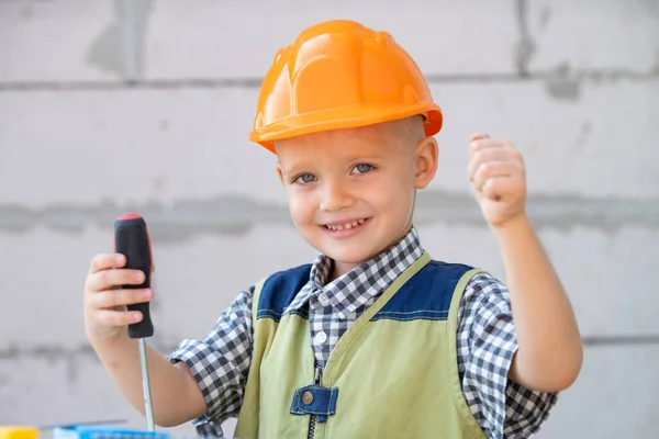 Happy smiling kid boy twists bolt with screwdriver. Child repairman with repair tool. Child in helmet and boilersuit on construction site. Little worker engineer. Kids builder and repair. — Stock Photo, Image