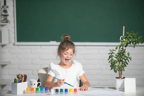 Dziecko dziewczyna rysuje w klasie siedzi przy stole, bawiąc się na tle tablicy szkolnej. Kreatywne kształcenie dzieci. — Zdjęcie stockowe