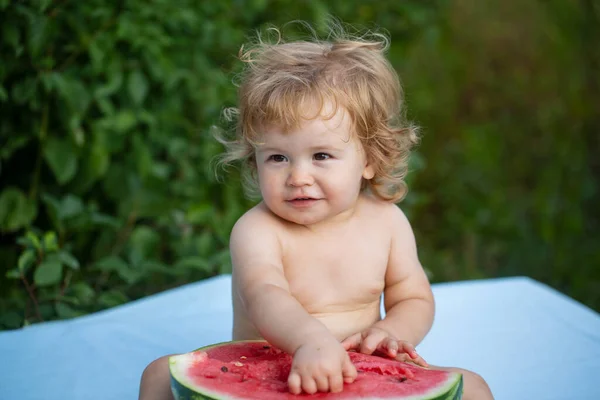 Bebé comendo melancia vermelha no jardim sentado na grama. Criança comendo melancia no jardim. As crianças comem fruta ao ar livre. Bebê bonito brincando no jardim segurando uma fatia de melancia. — Fotografia de Stock