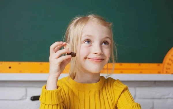 Közelkép egy csinos kis vidám kislányról, aki asztali asztalon ül az osztályteremben, csokoládéval. Kis vicces iskolás lány arc közelkép. — Stock Fotó