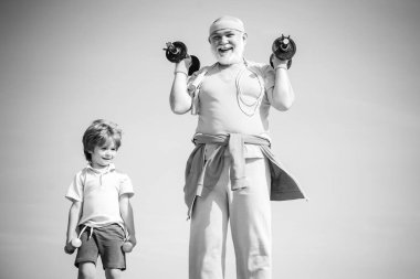 Kıdemli adam ve çocuk mavi gökyüzünde egzersiz yapıyorlar. Büyükbaba ve küçük torun halterleri üzerinde pratik yapıyorlar. Spor eğitimi.