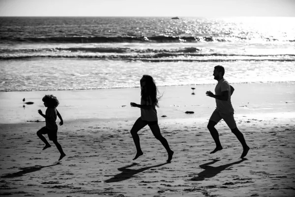 Los corredores de la familia se divierten en la playa correr y saltar al atardecer. Padre, madre, hijo pequeño corre. Niño saltar con diversión por la piscina de agua a lo largo del mar surf en la playa. —  Fotos de Stock