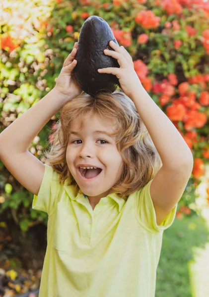 Söt pojke hålla avokado på huvudet, närbild porträtt på vid tropisk trädgård. — Stockfoto