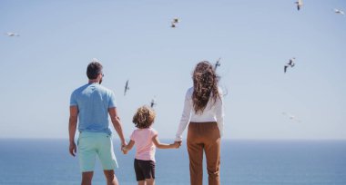Gökyüzündeki plajda aile manzarası. İnsanlar yaz tatilinde eğleniyor. Baba, anne ve çocuk mavi denize karşı el ele tutuşuyorlar. Tatil seyahati kavramı.