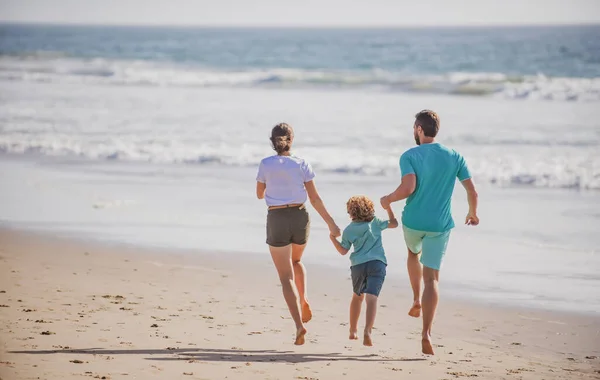 幸せな若い家族が走り、夏のビーチでジャンプします。家族のレジャー活動のコンセプト。親が走って飛び跳ねる子. — ストック写真