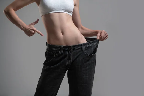 Dieting concept with oversized jeans. Skinny woman in too large jeans. Concept of successful weight loss. Thin girl in big trousers on a gray background. — стоковое фото