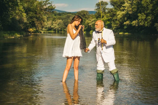 Giovane donna sexy e vecchio pescatore in piedi nel fiume con canna da pesca. Vecchio anziano e ragazza pesca. — Foto Stock