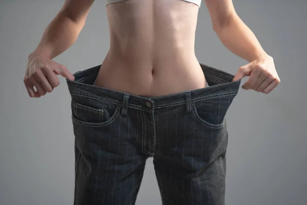 Cintura fina. A menina fina tem pontos na cintura fina em calças grandes, perda de peso bem sucedida, isolada no fundo cinza. Perda de peso bem sucedida, mulher com jeans muito grandes depois de uma dieta. — Fotografia de Stock