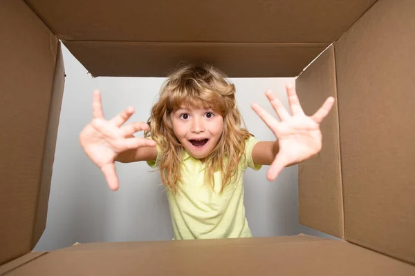 Child opens carton box and pulling out gift from it. Small boy looking in parcel box and happy to receiving a surprise. Young kid showing joy on his face. Unpacking cardboard box for kids. — стоковое фото