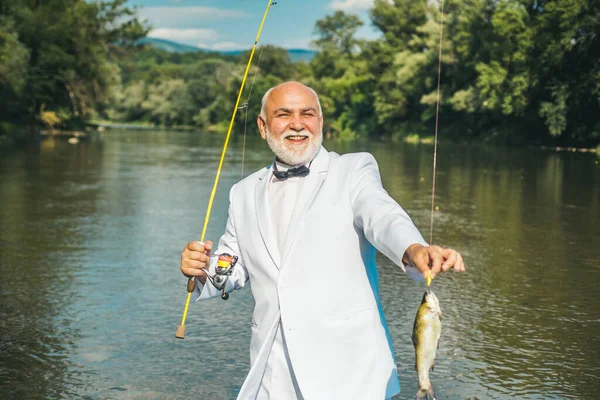 Un viejo pescador feliz pescó un pez trucha. Retrato de alegre sonriente hombre mayor pescando. Abuelo con pescado. Hombre maduro pescador en traje con caña de pescar, carrete giratorio en el río. — Foto de Stock