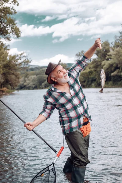 Pescatore utilizzando canna flyfishing nel fiume di montagna. Pescatore che pesca su un rever. Buon vecchio pescatore catturato un pesce trota. Ritratto di allegro sorridente uomo anziano pesca. Nonno con pescato. — Foto Stock