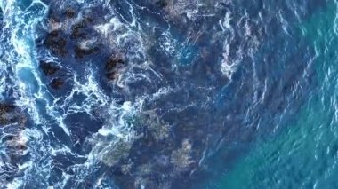 Dalgalar ve kayalar. Deniz dalgaları dokusu. İnsansız hava aracının doğa arkaplan görüntüsü. Köpüklü mavi okyanus suyu. Dalgalarda köpükle su sıçrıyor.