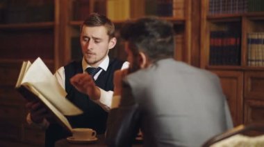 İş adamları kütüphanede sohbet ediyor. Erkekler konuşuyor ve tartışıyor. Klasik kütüphane dolabında kitap okuyan konsantre bir adam. Ev kütüphanesi. İş adamı klasik bir iç mekanda oturuyor, elinde kitap tutuyor..
